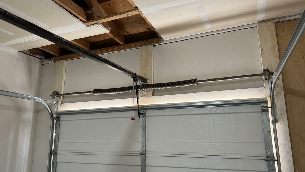 Interior of a tidy suburban garage looking up at a belt-drive opener rail and torsion spring above a closed insulated door, daylight leaking around the door edges